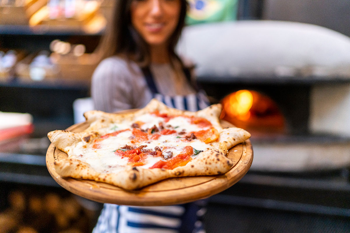 A smiling woman who works at a restaurant holds a freshly made pizza, reflecting the efficient service provided by the CAKE POS system.