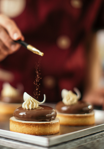 A baker's hand sprinkling powdered sugar on two chocolate pastries.