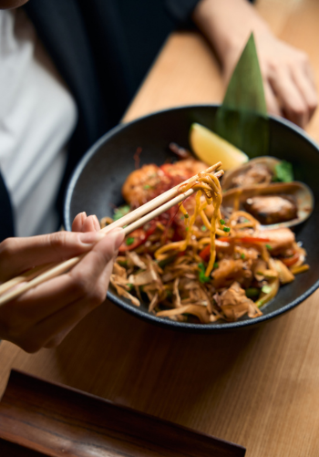 A person using chopsticks to eat a bowl of stir-fried noodles and shrimp.