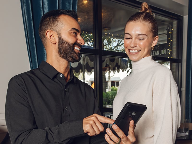 A man and a woman at a restaurant table look at a CAKEpop handheld POS, showing tableside ordering.