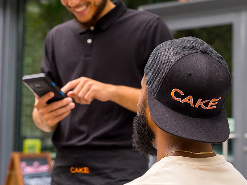 A waiter uses a CAKEpop handheld POS device to take an order from a customer who is wearing a backward-facing CAKE baseball hat.