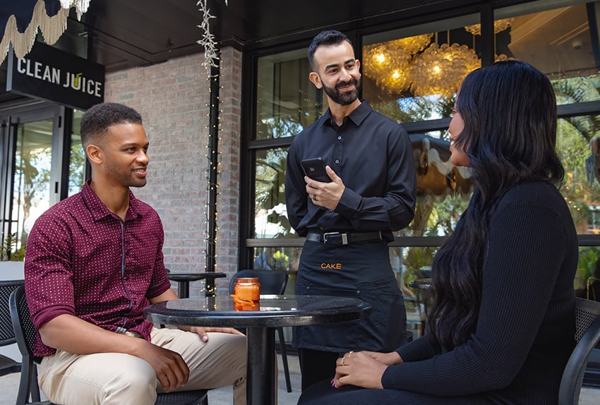 A waiter using the CAKEpop handheld POS device to take an order from two customers sitting at an outdoor table.