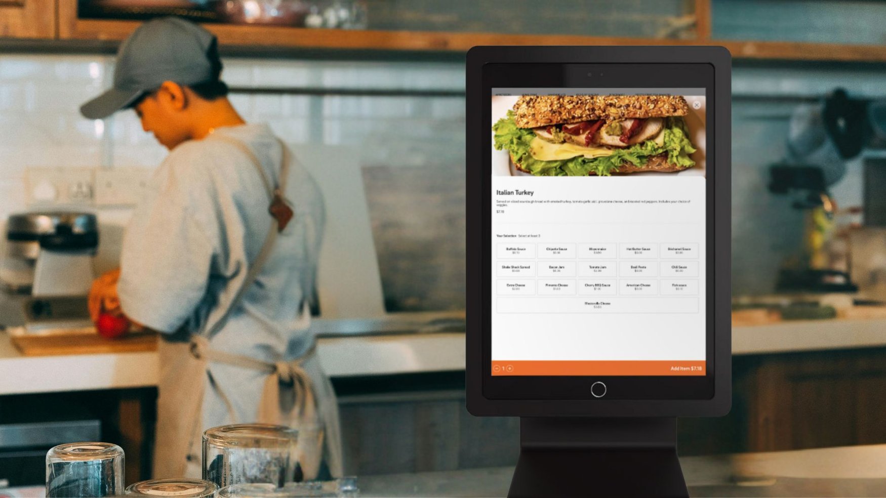 A customer using a CAKE Self-Service Kiosk in a fast-casual restaurant, with a restaurant employee in the background prepping an order